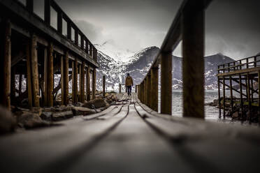 Ground level back view of explorer walking along wooden quay near sea in mountainous terrain in winter in Norway - ADSF21457