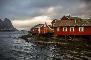 Rote Holzhäuser eines Fischerdorfs in der Nähe des Meeres in einer bergigen Gegend unter bewölktem Himmel in Norwegen - ADSF21462