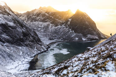 Entfernte Ansicht der Silhouette eines Wanderers, der auf einem verschneiten Hang in der Nähe des Meeres im Hochland steht, vor dem Hintergrund des Himmels bei Sonnenuntergang in Norwegen - ADSF21467