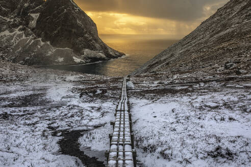 Wunderschöne Aussicht auf einen endlosen Wanderweg, der durch einen verschneiten Berghang zum Meer führt, unter dem Himmel des Sonnenuntergangs im Winter in Norwegen - ADSF21468