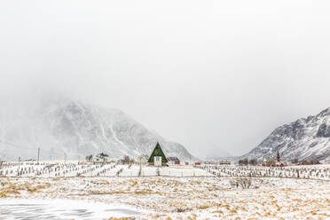 Landschaftliche Ansicht eines Holzhauses in der Nähe eines Friedhofs mit Grabsteinen in einem verschneiten Hochlandtal im Winter in Norwegen - ADSF21471