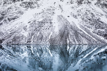 Berghang mit Schnee und blattlosen Bäumen, die sich in der glatten Wasseroberfläche eines Sees im Winter in Norwegen spiegeln - ADSF21475