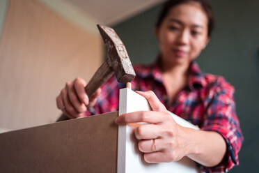 Fokussierte ethnische Frau, die einen Hammer zum Nageln von Holzdübeln benutzt, während sie neue Möbel in der Wohnung zusammenbaut - ADSF21510