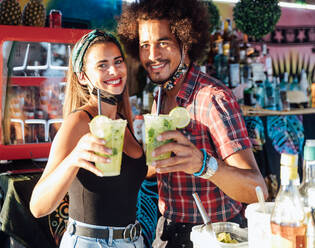 Positive ethnische Besitzer einer tropischen Bar, die freundlich in die Kamera schauen und ein Glas erfrischenden Cocktail mit Limette und Eis anbieten - ADSF21547