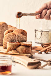 Crop unrecognizable person with dipper pouring sweet honey on fresh bread placed on cutting board on table in kitchen - ADSF21578