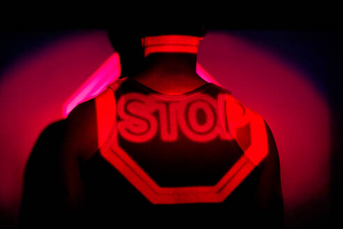Seitenansicht eines afroamerikanischen Mannes, der in einem dunklen Studio steht, beleuchtet von rotem Neonlicht der Projektion eines Stopp-Straßenschildes - ADSF21587