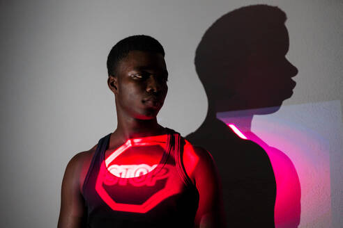 Emotionsloses afroamerikanisches männliches Modell, das in einem Studio mit roter Neonlichtprojektion eines Stopp-Straßenschilds steht, das das Konzept der Beschränkung zeigt - ADSF21588