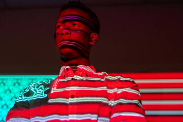 Serious African American male illuminated by red and blue neon projection of national American flag standing in dark studio - ADSF21590