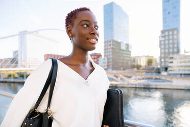 Selbstbewusste erfolgreiche positive junge schwarze Geschäftsfrau mit kurzer Frisur steht in der Nähe von Fluss gegen moderne Gebäude in der Innenstadt - ADSF21599