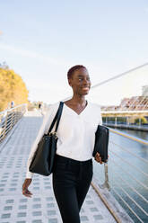 Selbstbewusste erfolgreiche positive junge schwarze Geschäftsfrau mit kurzer Frisur steht in der Nähe von Fluss gegen moderne Gebäude in der Innenstadt - ADSF21600
