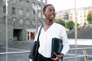 Confident successful positive young black businesswoman with short hairstyle standing near river against modern buildings in downtown - ADSF21601