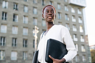 Niedrig angesetzte Seitenansicht einer selbstbewussten, lächelnden jungen afroamerikanischen Managerin in eleganter weißer Bluse, die eine Laptoptasche trägt und wegschaut, während sie in der Nähe eines modernen städtischen Gebäudes steht - ADSF21602