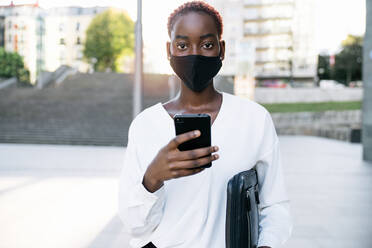 Stylish young African American businesswoman in protective mask standing on city street and messaging on smartphone - ADSF21604