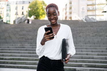 Junge afroamerikanische Geschäftsfrau in stilvollem Outfit liest eine Nachricht auf ihrem Smartphone und lächelt zufrieden, während sie auf einer Treppe in einer städtischen Straße steht - ADSF21605