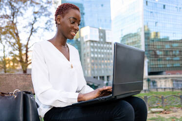 Positive junge afroamerikanische Geschäftsfrau in eleganter Kleidung sitzt auf einer Bank und arbeitet an einem Laptop vor einem unscharfen modernen städtischen Hintergrund - ADSF21608