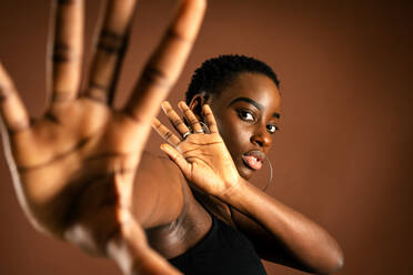 Black female model with short hair looking at camera and making protective gesture against brown background - ADSF21618