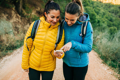 Fröhliche Wanderinnen mit Rucksäcken und in warmen Jacken stehen auf der Straße in der Natur und orientieren sich auf der GPS-Karte auf dem Smartphone während des Trekkings - ADSF21625