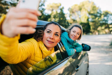 Fröhliche Freundinnen spähen aus dem Autofenster und machen zusammen ein Selfie mit dem Smartphone - ADSF21628