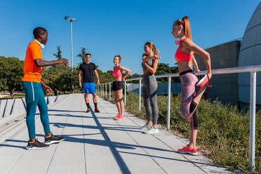 Group of multiethnic sportspeople stretching bodies while warming up before workout in city - ADSF21630