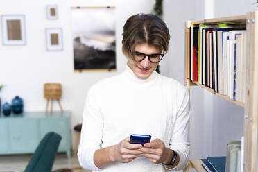 Junger Mann schreibt SMS über sein Smartphone, während er im Wohnzimmer neben einem Bücherregal steht - GIOF11736