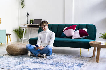 Junger Mann sitzt mit gekreuzten Beinen im Wohnzimmer und benutzt ein Smartphone - GIOF11740