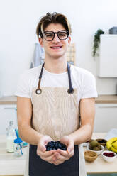 Smiling young man in apron holding fresh blueberries while standing in kitchen - GIOF11748