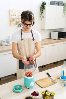 Young man putting chopped strawberries in blender while standing in kitchen - GIOF11753