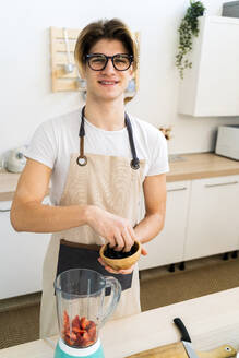 Smiling young man wearing eyeglasses with bowl of blueberries in kitchen - GIOF11754