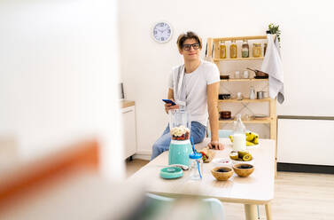 Junger Mann mit Smartphone sitzt auf dem Tisch in der Küche zu Hause - GIOF11764