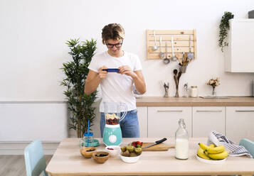 Junger Mann fotografiert Früchte im Mixer mit seinem Smartphone in der Küche zu Hause - GIOF11767