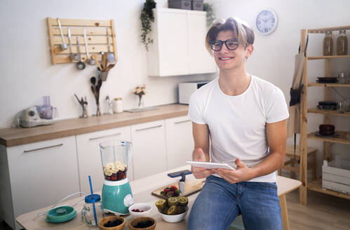 Smiling young man with digital tablet sitting on table in kitchen - GIOF11770