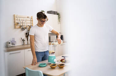 Man wearing eyeglasses pouring milk into blender while preparing smoothie in kitchen - GIOF11772