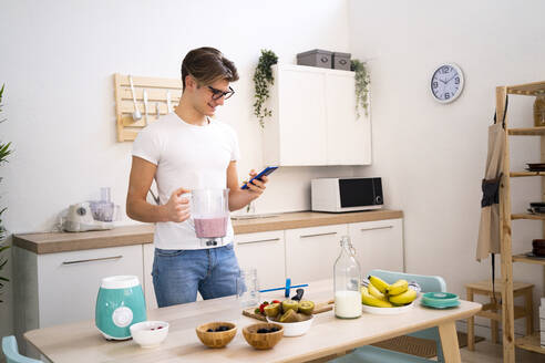 Young man holding blender of smoothie while using smart phone in kitchen - GIOF11777