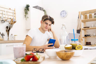 Young man using smart phone while sitting at table in kitchen - GIOF11791