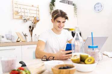 Smiling young man using smart phone while sitting at table in kitchen - GIOF11792
