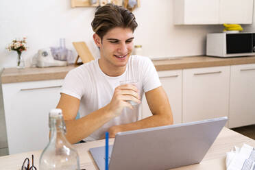 Junger Mann hält ein Glas Wasser in der Hand, während er zu Hause in der Küche am Laptop sitzt - GIOF11793