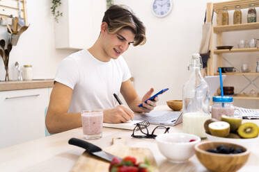 Young man with smart phone writing on notepad while sitting at table in kitchen - GIOF11795