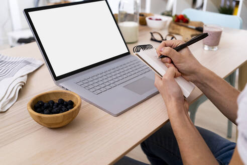 Mann sitzt mit Laptop am Tisch und schreibt auf einem Notizblock in der Küche zu Hause - GIOF11797