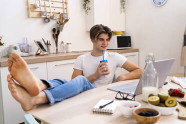 Relaxed young man drinking smoothie while sitting with laptop at table in kitchen - GIOF11799