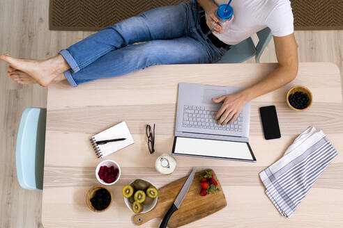 Mann hält Smoothie-Glas, während er am Tisch in der Küche sitzt und einen Laptop benutzt - GIOF11800