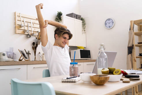 Young man stretching his hand while sitting at table with laptop in kitchen - GIOF11802