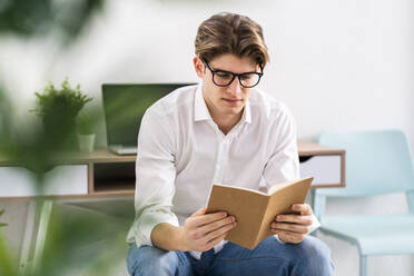 Junger Mann mit Brille liest ein Buch im Wohnzimmer - GIOF11810