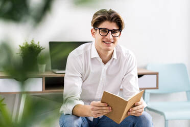 Lächelnder junger Mann mit Buch im Wohnzimmer sitzend - GIOF11811