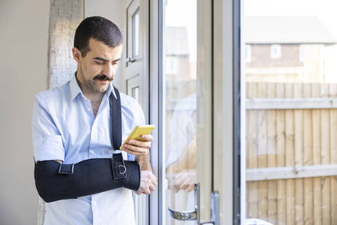 Mann mit Armschlinge, der sein Smartphone benutzt, während er zu Hause am Fenster steht - WPEF04217