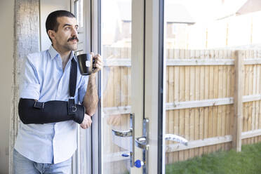 Thoughtful man wearing arm sling drinking tea while looking through window at home - WPEF04218