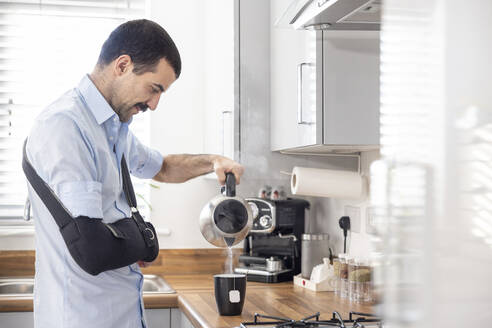 Injured man with arm sling pouring tea in cup through kettle in kitchen at home - WPEF04219