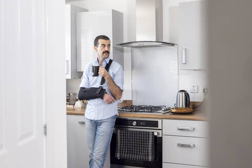 Man wearing arm sling while drinking tea in kitchen at home - WPEF04220