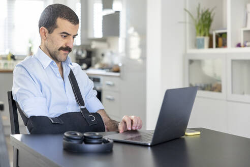 Mann mit Armschlinge arbeitet zu Hause am Laptop - WPEF04222