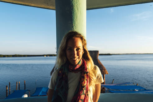 Lächelnde blonde Frau lehnt an einer architektonischen Säule gegen das Meer bei Sonnenuntergang - JMPF00880