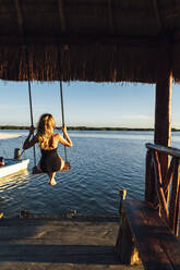 Mid adult woman swinging on swing in gazebo against sea during sunset - JMPF00882